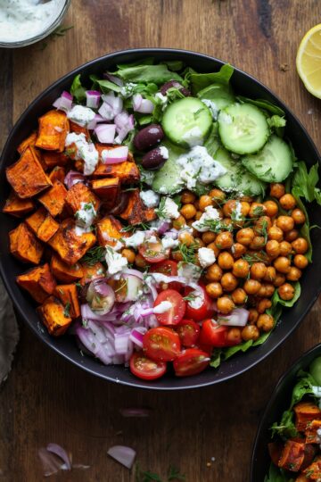 Overhead Greek chickpea sweet potato bowl with roasted sweet potatoes, spiced chickpeas, cucumber, cherry tomatoes, red onion, olives, leafy greens, feta, herbs, and creamy dressing in a black bowl.