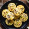 Overhead view of ham and mushroom egg bites topped with green onions on a black plate. The baked egg bites contain diced ham, mushrooms, and melted cheddar, surrounded by fresh mushrooms on a rustic wood background.