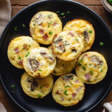 Overhead view of ham and mushroom egg bites topped with green onions on a black plate. The baked egg bites contain diced ham, mushrooms, and melted cheddar, surrounded by fresh mushrooms on a rustic wood background.