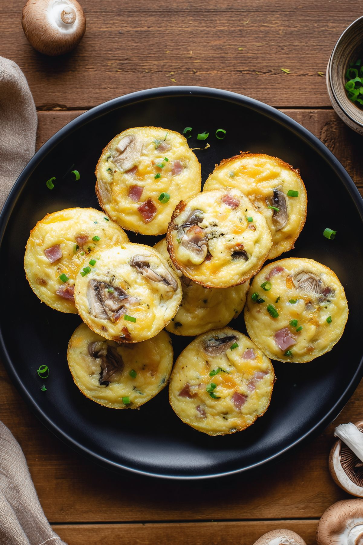 Overhead view of ham and mushroom egg bites topped with green onions on a black plate. The baked egg bites contain diced ham, mushrooms, and melted cheddar, surrounded by fresh mushrooms on a rustic wood background.