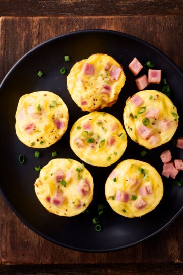 Six Ham and Swiss Egg Bites with diced ham, Swiss cheese, and green onions arranged on a matte black plate over a dark rustic wooden surface.
