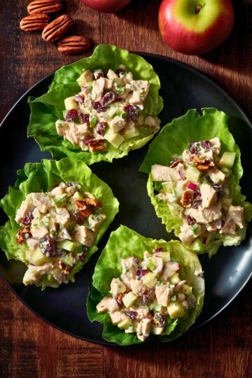 Overhead view of harvest chicken salad lettuce wraps with chicken, apples, dried cranberries, celery, and pecans on a black plate, with apples and pecans on the rustic wood table.