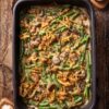Overhead view of healthy green bean casserole with fresh green beans, mushrooms, creamy sauce, and crispy fried onions in a black baking dish on a rustic wood table.