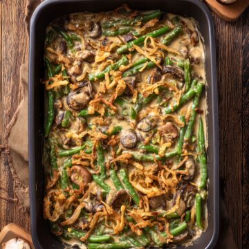 Overhead view of healthy green bean casserole with fresh green beans, mushrooms, creamy sauce, and crispy fried onions in a black baking dish on a rustic wood table.