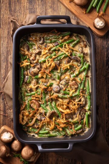 Overhead view of healthy green bean casserole with fresh green beans, mushrooms, creamy sauce, and crispy fried onions in a black baking dish on a rustic wood table.