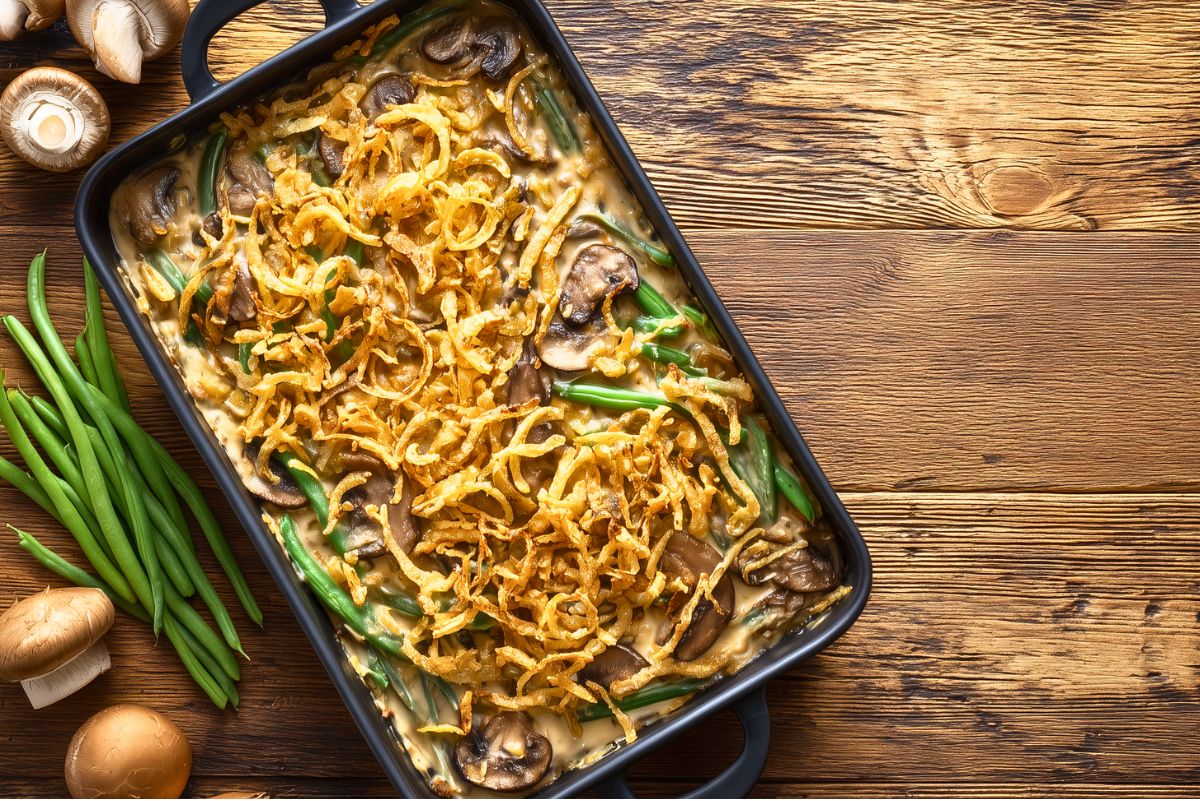 Healthy green bean casserole topped with golden crispy fried onions and mushrooms in a black baking dish, with fresh green beans and mushrooms on a rustic wood background.