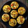 Overhead view of seven High Protein Taco Egg Bites with ground turkey, melted cheese, and red and green bell peppers arranged on a black plate over a dark rustic wood table.