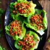 Overhead plate of hot honey chicken lettuce wraps with saucy chopped chicken, red bell pepper, carrots, and green onions in crisp lettuce leaves on a rustic wood table.