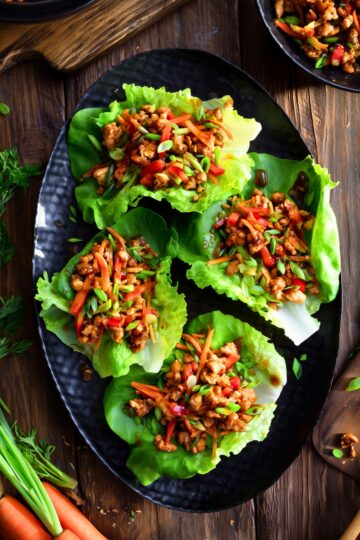 Overhead plate of hot honey chicken lettuce wraps with saucy chopped chicken, red bell pepper, carrots, and green onions in crisp lettuce leaves on a rustic wood table.