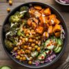 Overhead hot honey chickpea broccoli sweet potato bowl with roasted sweet potatoes, chickpeas, broccoli, cucumber, cabbage, red onion, creamy sauce, and hot honey drizzle in a black bowl.
