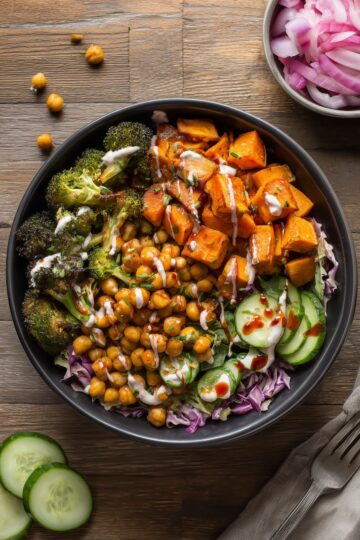 Overhead hot honey chickpea broccoli sweet potato bowl with roasted sweet potatoes, chickpeas, broccoli, cucumber, cabbage, red onion, creamy sauce, and hot honey drizzle in a black bowl.