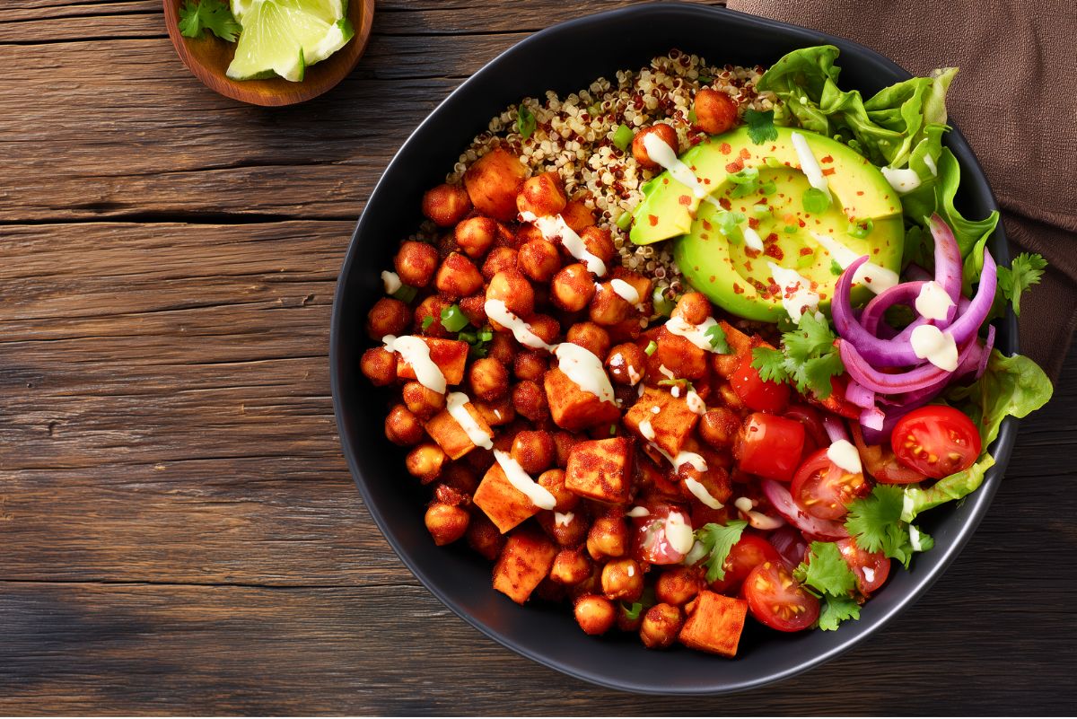 Hot Honey Chickpea Sweet Potato Bowl in a black bowl with roasted sweet potatoes, spicy chickpeas, quinoa, avocado, lettuce, tomatoes, red onion, cilantro, and creamy dressing on a rustic wood table.