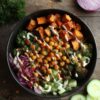 Hot Honey Chickpea Sweet Potato Bowl with roasted sweet potatoes, chickpeas, broccoli, cucumber, greens, red onion, herbs, and creamy sauce in a black bowl on a dark rustic wood table.