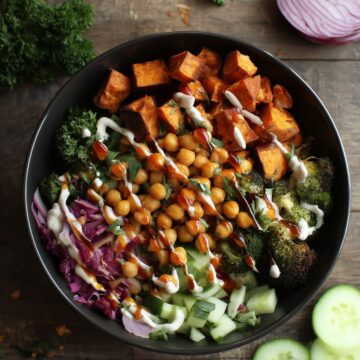 Hot Honey Chickpea Sweet Potato Bowl with roasted sweet potatoes, chickpeas, broccoli, cucumber, greens, red onion, herbs, and creamy sauce in a black bowl on a dark rustic wood table.