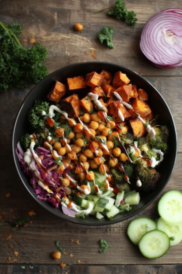 Hot Honey Chickpea Sweet Potato Bowl with roasted sweet potatoes, chickpeas, broccoli, cucumber, greens, red onion, herbs, and creamy sauce in a black bowl on a dark rustic wood table.