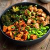 Hot honey garlic chicken sweet potato bowl with roasted sweet potatoes, broccoli, edamame, and green onions in a matte black bowl on rustic wood.