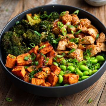 Hot honey garlic chicken sweet potato bowl with roasted sweet potatoes, broccoli, edamame, and green onions in a matte black bowl on rustic wood.