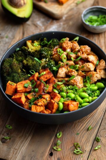 Hot honey garlic chicken sweet potato bowl with roasted sweet potatoes, broccoli, edamame, and green onions in a matte black bowl on rustic wood.
