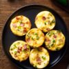 Six Jalapeño Popper Egg Bites with bacon, melted cheddar cheese, jalapeños, and green onions arranged on a matte black plate with fresh jalapeño peppers on a rustic wood background.