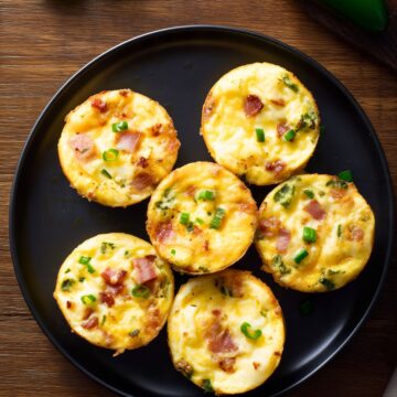 Six Jalapeño Popper Egg Bites with bacon, melted cheddar cheese, jalapeños, and green onions arranged on a matte black plate with fresh jalapeño peppers on a rustic wood background.