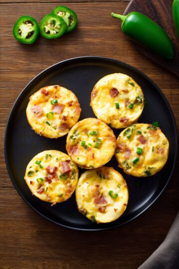 Six Jalapeño Popper Egg Bites with bacon, melted cheddar cheese, jalapeños, and green onions arranged on a matte black plate with fresh jalapeño peppers on a rustic wood background.