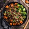 Overhead view of a Korean beef sweet potato bowl with seasoned ground beef, roasted cubed sweet potatoes, broccoli, green onions, and sesame seeds in a black bowl on a rustic wood background.