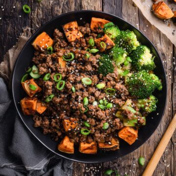 Overhead view of a Korean beef sweet potato bowl with seasoned ground beef, roasted cubed sweet potatoes, broccoli, green onions, and sesame seeds in a black bowl on a rustic wood background.