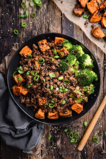 Overhead view of a Korean beef sweet potato bowl with seasoned ground beef, roasted cubed sweet potatoes, broccoli, green onions, and sesame seeds in a black bowl on a rustic wood background.