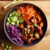 Korean chicken sweet potato bowl with glossy glazed chicken, roasted sweet potatoes, edamame, cucumber, shredded carrots, purple cabbage, green onions, and sesame seeds in a black bowl.