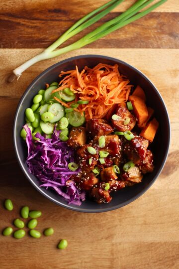 Korean chicken sweet potato bowl with glossy glazed chicken, roasted sweet potatoes, edamame, cucumber, shredded carrots, purple cabbage, green onions, and sesame seeds in a black bowl.