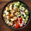 Overhead lemon garlic chicken protein bowl with quinoa, seasoned chicken, broccoli, cucumber, red bell pepper, red onion, herbs, and creamy dressing in a black bowl on a rustic wooden table.