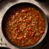 Overhead view of Lentil Soup Vincent in a black bowl, filled with lentils, carrots, celery, onions, and tomatoes in a savory broth, styled on a rustic surface with fresh vegetables nearby.