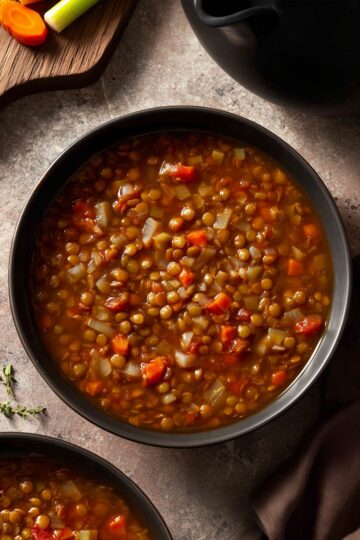 Overhead view of Lentil Soup Vincent in a black bowl, filled with lentils, carrots, celery, onions, and tomatoes in a savory broth, styled on a rustic surface with fresh vegetables nearby.