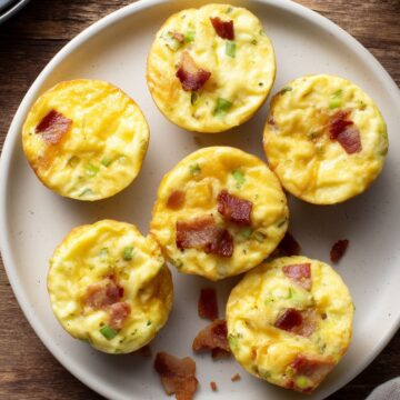 Six maple bacon egg bites with cheddar, crispy bacon, and green onions arranged on a white plate over a rustic wood table.