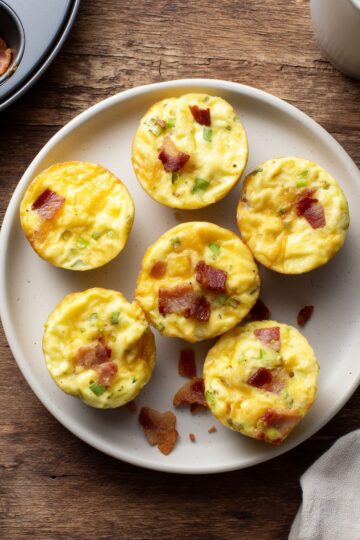Six maple bacon egg bites with cheddar, crispy bacon, and green onions arranged on a white plate over a rustic wood table.