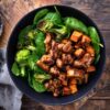 Overhead view of a maple balsamic chicken sweet potato bowl with glazed chicken, roasted sweet potatoes, broccoli, and spinach in a black bowl on a rustic wooden board.