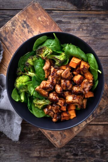 Overhead view of a maple balsamic chicken sweet potato bowl with glazed chicken, roasted sweet potatoes, broccoli, and spinach in a black bowl on a rustic wooden board.