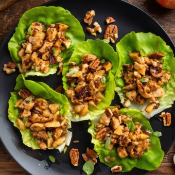 Overhead view of maple glazed chicken and pecan lettuce wraps on a black plate, with chopped apples, pecans, and green onions. Fresh apple and scattered pecans sit on the rustic wood table.