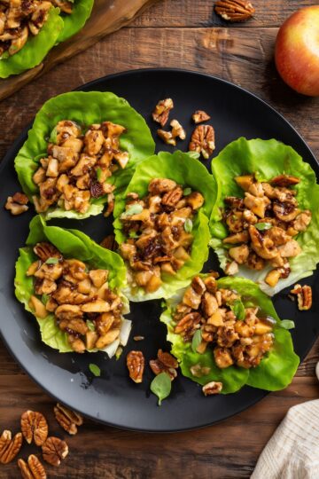 Overhead view of maple glazed chicken and pecan lettuce wraps on a black plate, with chopped apples, pecans, and green onions. Fresh apple and scattered pecans sit on the rustic wood table.