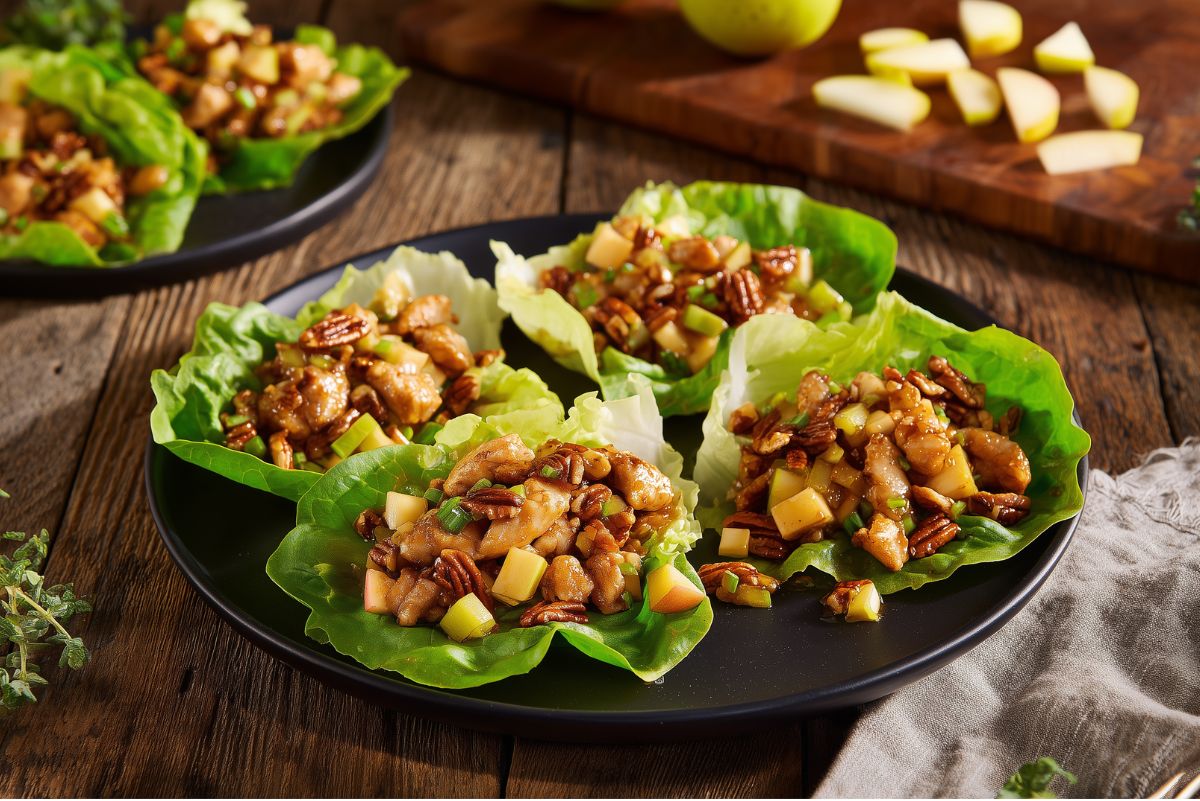 Maple glazed chicken and pecan lettuce wraps arranged on a black plate with diced chicken, apples, pecans, and green onions. A cutting board with sliced apples sits in the background on a rustic wood table.