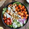 Overhead view of a Mediterranean chicken sweet potato protein bowl with roasted sweet potatoes, chicken, chickpeas, cherry tomatoes, cucumber, red onion, feta, greens, and creamy herb dressing in a black bowl on rustic wood.
