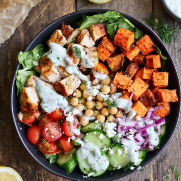 Overhead view of a Mediterranean chicken sweet potato protein bowl with roasted sweet potatoes, chicken, chickpeas, cherry tomatoes, cucumber, red onion, feta, greens, and creamy herb dressing in a black bowl on rustic wood.