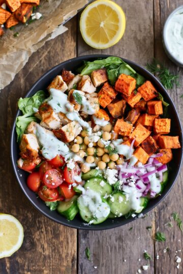 Overhead view of a Mediterranean chicken sweet potato protein bowl with roasted sweet potatoes, chicken, chickpeas, cherry tomatoes, cucumber, red onion, feta, greens, and creamy herb dressing in a black bowl on rustic wood.
