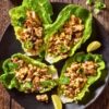 Overhead view of four Mexican Street Corn Chicken Lettuce Wraps filled with grilled chicken, corn, red onion, jalapeño, crumbled cheese and cilantro on a black plate over a rustic wood table.
