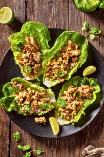 Overhead view of four Mexican Street Corn Chicken Lettuce Wraps filled with grilled chicken, corn, red onion, jalapeño, crumbled cheese and cilantro on a black plate over a rustic wood table.