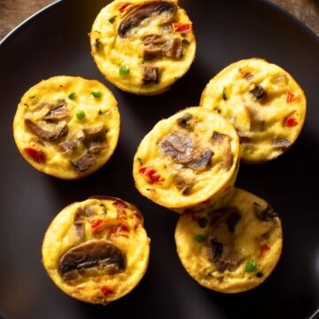 Mushroom and roasted red pepper egg bites with browned mushrooms, red pepper pieces, and green onion on a matte black plate.