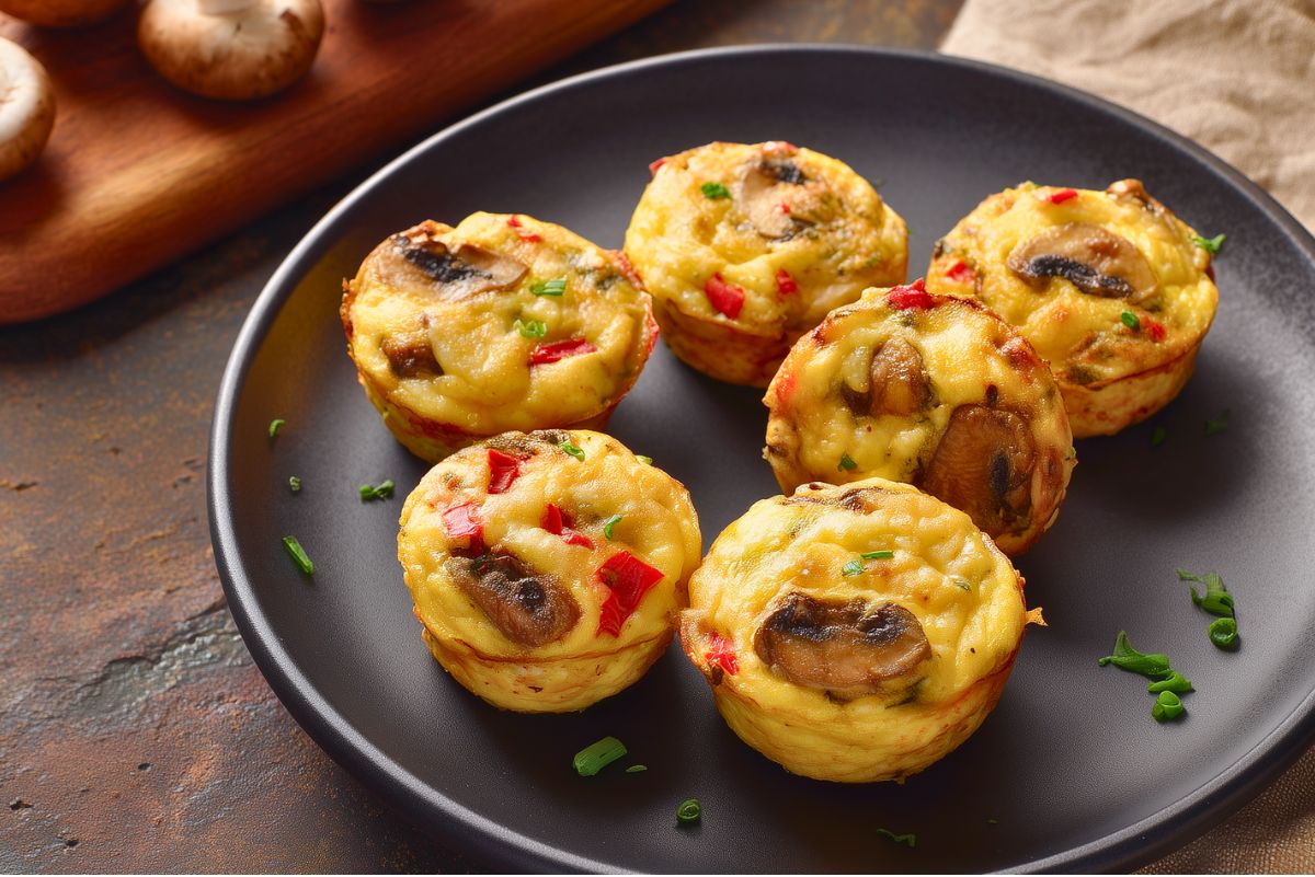Six mushroom and roasted red pepper egg bites with golden edges, sliced mushrooms, red peppers, and green onion on a dark plate.