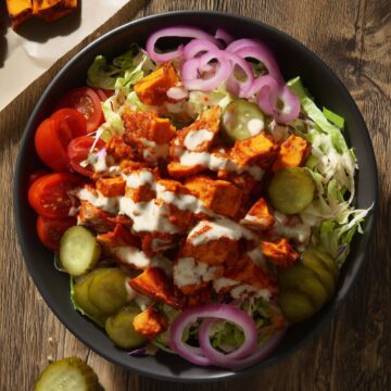 Overhead Nashville hot chicken sweet potato bowl with seasoned chicken, roasted sweet potatoes, cabbage, cucumbers, tomatoes, red onion, pickles, and creamy dressing in a black bowl.