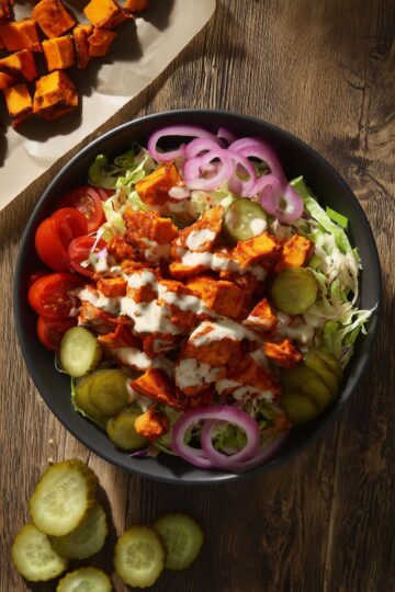 Overhead Nashville hot chicken sweet potato bowl with seasoned chicken, roasted sweet potatoes, cabbage, cucumbers, tomatoes, red onion, pickles, and creamy dressing in a black bowl.