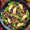 Overhead view of pear pecan fall salad with mixed greens, sliced pears, dried cranberries, toasted pecans, crumbled feta, and red onion in a black bowl on a rustic wood table.